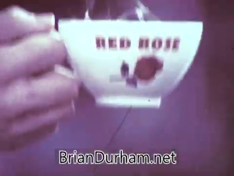 The still features a close-up of a hand holding a cup labeled 'Red Rose,' showcasing the brand prominently. The cup likely emphasizes a nostalgic aesthetic typical of the late 1960s. The background appears slightly blurred, drawing focus to the cup itself, which suggests a tea-drinking moment, possibly in an inviting or cozy setting. The overall tone reflects the advertising style of the time, aiming to connect with viewers through warmth and familiarity.
