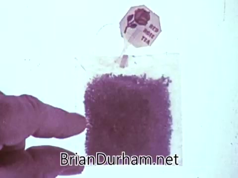 The still from the 1968 'Another Red Rose Tea' commercial features a hand pointing towards a teabag filled with loose tea leaves. The teabag has a tag labeled 'Red Rose Tea,' showcasing the product prominently. The background is light, drawing focus to the teabag and the hand, effectively highlighting the tea's quality. This image captures a moment intended to promote the brand's appeal in a straightforward manner.