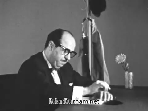 The still from the 1968 PSA 'Get yourself tested for diabetes' features a middle-aged man, likely portraying a medical professional, sitting at a desk. He appears to be engaging with a patient, emphasizing the importance of diabetes testing for individuals over 40, particularly if they show signs like fatigue or have a family history of the condition. The setting is simple, with minimal props—a coat, a hat, and a vase with a flower—underscoring the seriousness of the health message.
