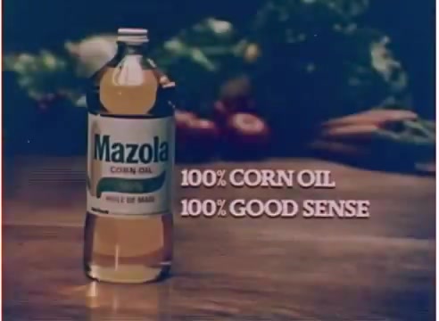 The still features a bottle of Mazola corn oil prominently displayed against a backdrop that suggests fresh produce, typical of 1970s food advertisements. The text highlights '100% CORN OIL' and '100% GOOD SENSE,' emphasizing the product's purity and health aspects. The overall aesthetic is nostalgic, reflecting the advertising style of the era.