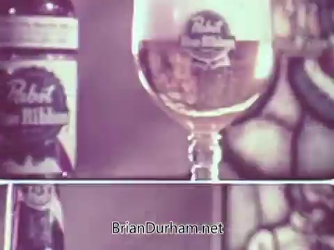 The still from the Pabst Beer short commercial features a close-up of a bottle and a glass of Pabst beer. The bottle is prominently labeled with the Pabst logo, and the glass displays the beer's golden color, highlighting its refreshing qualities. The background suggests a cozy or vintage setting, enhancing the nostalgic appeal of the advertisement. The text 'at popular prices' emphasizes affordability, targeting a broad audience.