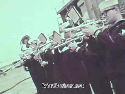 The still depicts a group of sailors in military uniforms playing brass instruments, likely trumpets, as part of a public service announcement for the US Navy from 1968. The sailors are gathered in a formation, suggesting a ceremonial context, with a backdrop that indicates an outdoor setting. The image captures a moment of patriotic performance.