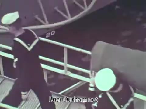 The still features two Navy sailors in uniform walking on a gangway. One sailor is carrying a cylindrical object, while the other is walking ahead. The setting appears to be near a ship, with a boat hull in the background. The image captures a moment of daily naval life from 1968, emphasizing camaraderie and activity within the Navy.