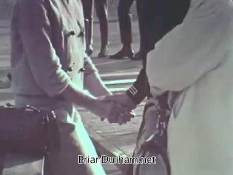 The still from the 1968 US Navy PSA features two people holding hands, emphasizing a moment of connection or support. The setting appears informal, with figures in the background, suggesting a public or community space. The focus on the hands implies themes of unity, friendship, or camaraderie, likely related to the larger message of the PSA promoting the Navy or military service.