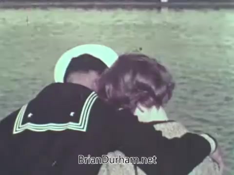 The still shows a close-up of a sailor in uniform, featuring a white hat with a black band, embracing a woman. They are positioned closely together, with the focus on their affectionate connection. The background features water, creating a serene and nostalgic atmosphere, typical of mid-20th century public service announcements promoting the Navy.