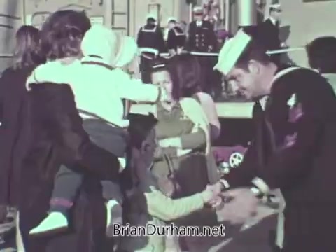 The still depicts a scene from a 1968 US Navy public service announcement. It shows a sailor interacting with children and families, likely at a naval base or welcome event. The environment appears joyful, emphasizing community and familial connections, as the sailor is engaging warmly with the kids. The setting features naval personnel in the background, highlighting the connection between the military and family life.
