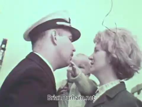 The still depicts a tense moment between a man in a Navy officer's uniform and a woman, possibly his partner, as they share a close interaction. A child is present, reaching towards the officer, suggesting a family dynamic. The scene is set against a light greenish backdrop, typical of the 1960s aesthetic in public service announcements.
