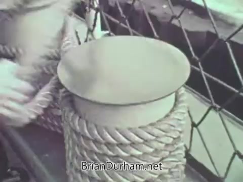 The 16mm still from 'US Navy PSA - 1968' features a close-up of a mooring bollard topped with a circular cap, surrounded by thick, twisted ropes. A hand is seen interacting with the equipment, indicative of tie-up procedures on a naval vessel. The setting suggests a maritime environment, emphasizing the operational tasks and safety protocols associated with navy activities.