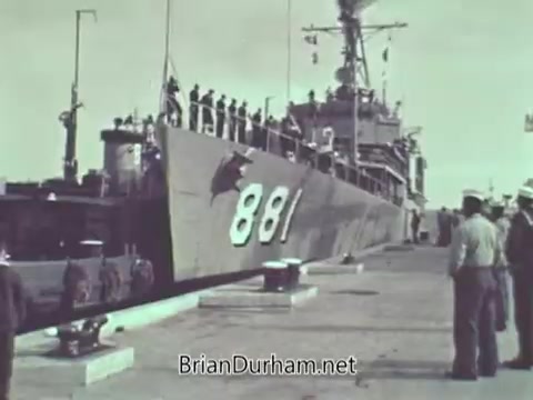 The still features a US Navy ship, labeled with the number '881,' docked at a port. Sailors and personnel are visible on the ship and around the dock, suggesting an active naval environment. The overall scene conveys military life and operations during the late 1960s.