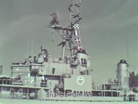 The still depicts a naval ship from the US Navy, showcasing its deck and equipment. Various flags are displayed, including signal flags on the mast, indicating communication capability. The ship appears to be in an operational setting, reflecting the military's focus on readiness during the late 1960s. The image is in black and white, typical of that era's media.