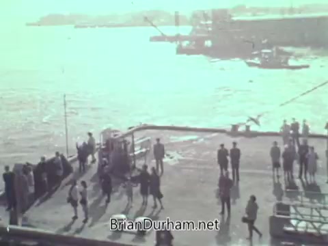 The still from 'US Navy PSA - 1968' depicts a busy waterfront scene with groups of people on a pier. Some individuals appear to be interacting or socializing, while others watch a boat on the water. The atmosphere suggests activity and engagement around naval life, with sunlight reflecting off the water, adding a warm tone to the setting.
