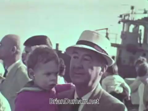 The still shows a man wearing a light-colored suit and hat holding a young child. They are in a crowd by the water, with a boat visible in the background. The scene appears to capture a moment of family or community gathering, likely related to a Navy event, given the context of the PSA. The overall tone is nostalgic and focused on human connection.