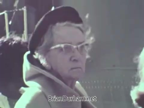 The still from the 1968 US Navy PSA features an elderly woman wearing glasses and a black beret, looking intently at something off-camera. She is dressed in a light-colored coat, suggesting a public setting. The background appears blurred, focusing attention on her expression, which conveys seriousness or contemplation.