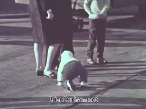 The still from 'US Navy PSA - 1968' features a child crawling on the ground in a public space. Adults are nearby, suggesting a familial or communal setting. The scene likely aims to convey themes of safety or community engagement, typical for a public service announcement. The overall tone appears candid and relatable, focusing on everyday life.