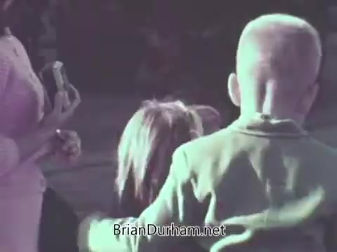 The still from the 1968 US Navy PSA depicts a group of children, with one child prominently in the foreground wearing a light-colored sweater and another child behind them. An adult, presumably a parent, is holding a camera and appears to be taking a photograph. The scene conveys a sense of innocence and familial connection. The colors suggest a vintage aesthetic typical of that era in filmmaking.