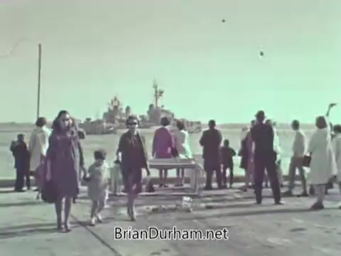 The still depicts a group of people on a dock, with a naval ship in the background. Some individuals are engaged in playful activities, while others are diverse in age and clothing, suggesting a casual, community atmosphere. The scene appears to be from the late 1960s, likely promoting the US Navy's connection with the public and family engagement.