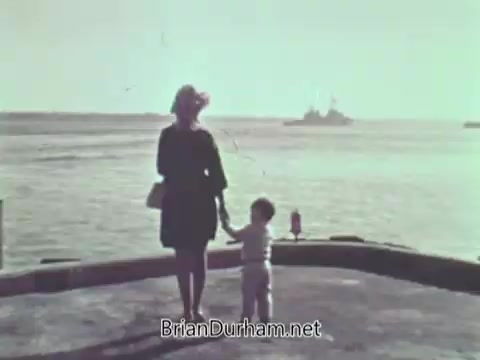 The still features a woman and a young child standing on a dock, gazing out toward the water. In the background, a naval ship is visible in the distance, indicating a coastal setting. The image has a nostalgic, vintage quality, typical of the late 1960s. The mood suggests themes of separation or longing, which are common in public service announcements depicting military families.