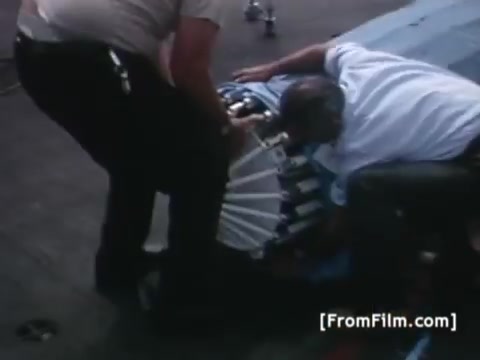 The still depicts personnel working on a piece of equipment, likely related to satellite communication, on the deck of the USS Hornet. Two individuals are engaged in a hands-on task, focusing on setting up or repairing a device used for transmitting images, particularly associated with the Apollo 11 recovery. The scene captures the high-stakes environment surrounding the historic mission and the collaboration necessary for successful satellite operations.