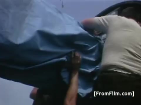 The still captures a scene of individuals preparing equipment for the Apollo 11 mission, specifically related to Western Union International. Workers are seen lifting a large blue tarp or cover, likely concealing satellite technology intended to send images from the USS Hornet. The focus on teamwork and logistics highlights the effort involved in post-mission recovery and communication operations.