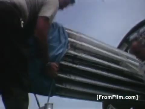 The still captures a moment during the preparation for the Apollo 11 mission, showing a worker handling equipment covered with a blue tarp. The focus is on the physical labor involved in setting up communication technology, likely antennas or satellite components, highlighting the logistical efforts by Western Union International to transmit satellite images of the recovery operation from the USS Hornet. The scene reflects the urgency and importance of communications in the context of the historic moon landing.