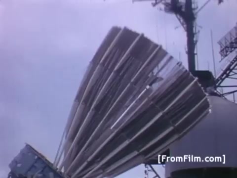 The still depicts a large satellite dish or antenna pointed skyward, likely part of the communication equipment used by Western Union International. It showcases the preparations for transmitting images of the Apollo 11 recovery operation via the USS Hornet, highlighting the technological setup essential for real-time communication during the historic mission. The scene is characterized by a cloudy sky, suggesting the operational conditions during the launch or recovery process.