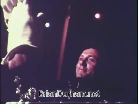 The still shows a smiling man looking up as a hand, possibly offering food, is raised towards him. The scene is warm and inviting, with soft lighting creating an homey atmosphere. It appears to capture a moment of interaction, likely highlighting the appeal of the dog food being advertised.