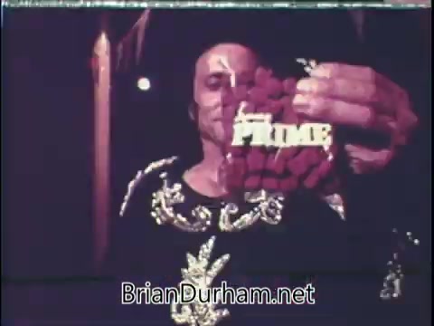 The still features a man holding a bag of 'Prime' dog food, prominently displayed in front of him. He appears to be in a casual setting, possibly outdoors or in a dimly lit area, wearing a decorative outfit. The focus is on the dog food packaging, highlighting the brand name. The overall aesthetic is characteristic of vintage advertising.