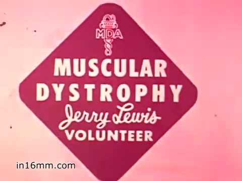 The still from the 1960s Muscular Dystrophy animated PSA features a bold diamond-shaped graphic with the text 'MUSCULAR DYSTROPHY' prominently displayed. Below it, the words 'Jerry Lewis VOLUNTEER' are written in a playful script. The background is a soft pink hue, enhancing the visibility of the text and giving it a warm, inviting feel. The overall design conveys a sense of urgency and community support, encouraging donations and awareness for the cause.