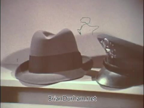The still from the 'Air Force Reserve recruiting PSA' features two hats prominently displayed: a civilian-style fedora and a military officer's cap. This visual juxtaposition symbolizes the blend of civilian life and military service, emphasizing the recruiting campaign targeting doctors during the Vietnam War era. The setting appears simple, highlighting the hats against a neutral background, suggesting a call to action for medical professionals to serve.