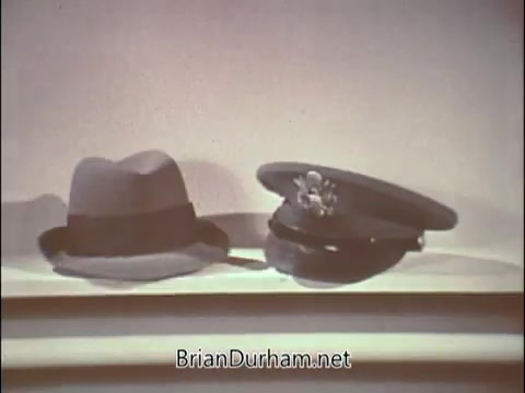 The still features two hats on a simple background: a civilian fedora and a military officer's cap, highlighting the juxtaposition between civilian life and military service. This imagery is typical of recruitment efforts from the Vietnam War era, specifically aimed at physicians to encourage their enlistment in the Air Force Reserve. The visual suggests a transition from civilian roles to military responsibilities.