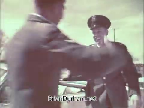 The still from the 'Air Force Reserve recruiting PSA' features a scene where a uniformed Air Force officer greets another man, likely a doctor. The setting appears to be outdoors, possibly near vehicles, reflecting the mid-20th century style of the era. The image conveys a sense of camaraderie and professionalism, characteristic of recruitment efforts during the Vietnam War period, targeting medical professionals to join the reserves.