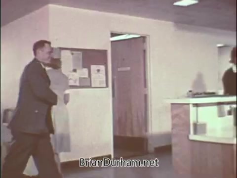 The still portrays a scene from a 1960s Air Force Reserve recruiting PSA targeting doctors. It features a man in a military uniform walking through a hospital corridor, suggesting a professional medical environment. The backdrop includes bulletin boards and a reception area, evoking a sense of urgency and duty, typical of military recruitment pieces during the Vietnam War era. The overall tone reflects the blending of medicine and military service, appealing to doctors to join the reserves.