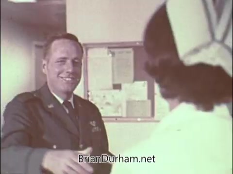 The still shows a man in an Air Force uniform smiling and extending his hand in a handshake to a woman, likely a nurse, who is wearing a medical outfit. The setting appears to be a hospital or clinic, indicated by the background elements like bulletin boards. This PSA from the 1960s is aimed at recruiting doctors into the Air Force Reserve, reflecting the era's military and medical themes during the Vietnam War. The tone is likely intended to convey camaraderie and a sense of service.