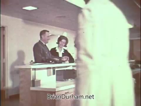 The still from the Air Force Reserve recruiting PSA features a modern, bright hospital or clinic setting. A man in military uniform converses with a woman dressed in professional attire at a reception desk. In the foreground, a doctor in a white coat walks by, suggesting a busy medical environment. The scene conveys an impression of professionalism and teamwork, aimed at attracting doctors to serve in the Air Force Reserve during the Vietnam War era. The overall tone is optimistic, highlighting opportunities for medical professionals.