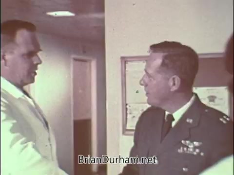 The still shows a handshake between two men in a professional setting. One is in a white lab coat, likely a doctor, while the other is in an Air Force uniform, indicating a recruitment context. This scene reflects a 1960s Air Force Reserve PSA aimed at attracting medical professionals during the Vietnam War era. The setting suggests a hospital or clinic environment, emphasizing the military's need for skilled doctors.