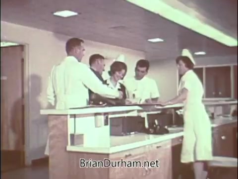 The still depicts a scene in a medical setting, likely a hospital or clinic, featuring a group of healthcare professionals. There are four individuals in white coats, including one doctor, and a nurse in uniform, gathered around a counter, seemingly discussing or reviewing information. The environment is bright and clinical, reflecting the era's aesthetic. This 1960s PSA aims to recruit medical professionals for the Air Force Reserve during the Vietnam War period, highlighting the importance of doctors in military service.