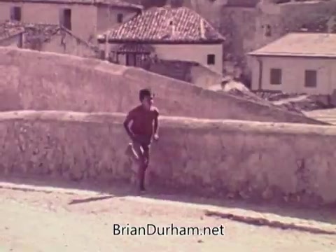 The still from the 1972 Coca-Cola commercial depicts a young athlete running along a rural path, with a backdrop of rustic buildings and a tiled roof. The scene conveys a sense of determination and energy, likely intended to evoke the spirit of the Olympic Games, emphasizing themes of youth and vitality amidst the contrasting serene environment. The context of being linked to the 1972 Munich Olympics adds a layer of poignancy to the imagery.