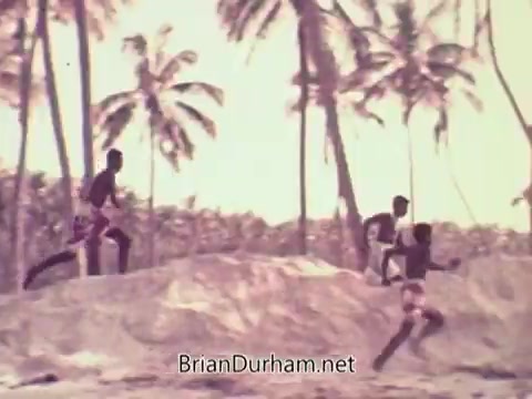 The still from the 1972 Coca-Cola commercial depicts a group of young boys running on a sandy beach, framed by palm trees. The scene conveys a sense of playfulness and vitality, representing the spirit of youth and athleticism associated with the Olympic games. The warm tones and vintage quality of the footage evoke a nostalgic atmosphere, contrasting with the serious backdrop of the Munich Olympics that year.