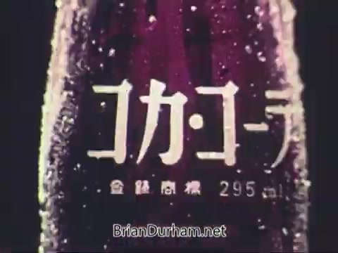 The still features a close-up of a Coca-Cola bottle, prominently displaying the brand name in stylized Japanese characters. The image captures the iconic shape of the bottle with condensation, emphasizing refreshment. The background is blurred, putting focus on the bottle, likely aiming to evoke a sense of enjoyment and connection during the 1972 Munich Olympics, despite the event's overshadowing tragedy.