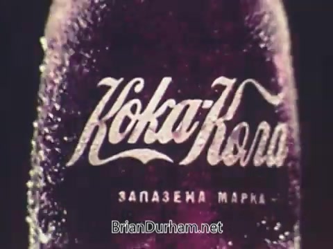 The still from the 1972 Olympics Coca-Cola commercial features a close-up of a Coca-Cola bottle, prominently displaying the iconic script logo. The bottle is glistening with condensation, indicating refreshment and appeal. The backdrop likely enhances the emphasis on the beverage, capturing the spirit of the Olympic Games, though overshadowed by the tragic events in Munich. The overall aesthetic evokes a sense of nostalgia and highlights Coca-Cola's branding during a significant historical moment.