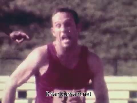 The still captures a close-up of a male athlete in a red tank top, running with intense focus. His expression shows a mix of determination and strain, reflecting the competitive spirit of the Olympics. The background is blurred, emphasizing the athlete's movement, while the overall vintage look of the image reflects the era of the 1972 Munich Olympics. The context of the commercial connects to the themes of sportsmanship and camaraderie, set against the backdrop of a tense historical event.