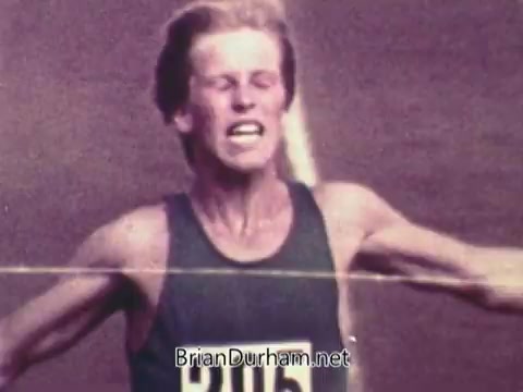The still from the 1972 Coca-Cola commercial features a male athlete with a pained expression, emphasizing the intense emotions of competition. He is wearing a tank top and has a race number displayed prominently on his chest. The image captures a moment of effort, likely as he nears the finish line, illustrating the spirit of determination and athleticism associated with the Olympics, set against the backdrop of the Munich Games, which were overshadowed by tragedy.