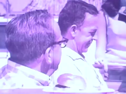 The still features two men enjoying a casual moment outdoors, likely in a social setting. One wears glasses and a checked shirt, while the other has a lighter-colored shirt and a pleasant expression. The background appears bright and warm, indicative of a sunny day, and hints at a lively atmosphere typical of a commercial. The overall color tone has a vintage quality, characteristic of 16mm film.