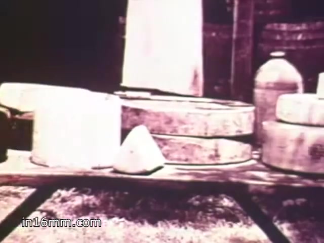 The still from 'Mueller's Elbows' shows a rustic scene featuring several large cheese wheels and a couple of jars or containers arranged on a wooden table. The environment appears outdoor, possibly in a farm setting, highlighting a vintage aesthetic typical of 1970s commercials. The monochromatic tones give it a classic look, emphasizing the products on display.