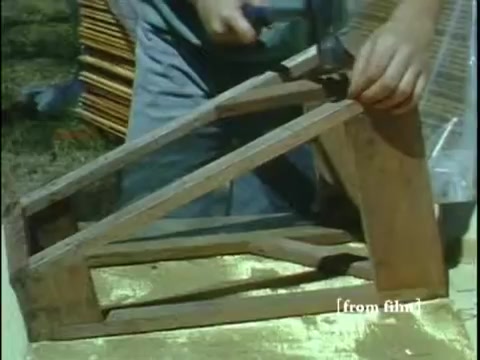 The still from 'Collecting Large Bottom Dwelling Organisms' shows a person constructing a wooden frame, likely a tool or device used for collecting aquatic organisms. The hands are actively engaged in manipulating the wood, with attention to detail on the framing. The surrounding environment suggests an outdoor setting, likely related to the film's theme of biodiversity exploration. The film is characterized by its vibrant Ektachrome color palette typical of the late 1960s.
