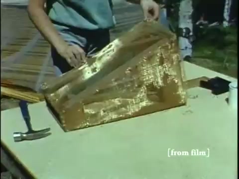 The still from 'Collecting Large Bottom Dwelling Organisms' depicts a person preparing equipment on a table. The focus is on a shiny, golden-brown box, possibly an aquatic trap, being handled carefully. Tools like a hammer and other supplies are visible, suggesting a hands-on approach to collecting organisms. The scene likely captures a moment of preparation for fieldwork, highlighting the practical aspects of gathering small frogs and other creatures from underwater environments.