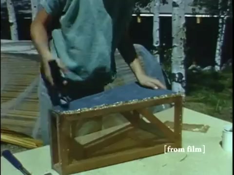 The still from 'Collecting Large Bottom Dwelling Organisms' depicts a person in a blue shirt working on a wooden structure at a table. The structure appears to be a container, possibly for housing small frogs and other creatures, with a mesh top. The setting includes trees in the background, indicating an outdoor environment. The image captures a moment of hands-on crafting, likely related to the preparation for collecting specimens. The film quality suggests a vibrant color palette typical of late 1960s Ektachrome.