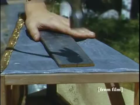 The still from 'Collecting Large Bottom Dwelling Organisms' depicts a hand hammering a wooden plank on a workbench. The setting appears outdoors, with hints of nature visible in the background. This scene likely illustrates preparations for collecting small aquatic creatures, such as frogs, highlighting the hands-on, instructional nature of the film from the late 1960s. The image captures a moment of craftsmanship involved in the process.