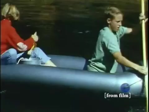 The still features two children on an inflatable raft in a body of water, likely illustrating the process of collecting small bottom-dwelling organisms like frogs. One child is seated and appears to be preparing to collect specimens, while the other is rowing. The scene captures a serene, educational moment from a late 1960s film focusing on nature and exploration. The vibrant colors typical of Ektachrome film enhance the lively atmosphere of the outdoor setting.