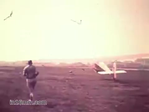 The still from the 1960s US Army recruiting PSA 'Your Man in Nürnberg' depicts a soldier, presumably Joe Naifus, running across an open field towards a small airplane. The scene captures a serene yet busy atmosphere, emphasizing both the soldier's dedication and the operational complexities of military logistics. In the background, birds fly, contributing to the tranquil environment of Nuremberg, while the soldier embodies the message of competence and adventure offered by the Army.